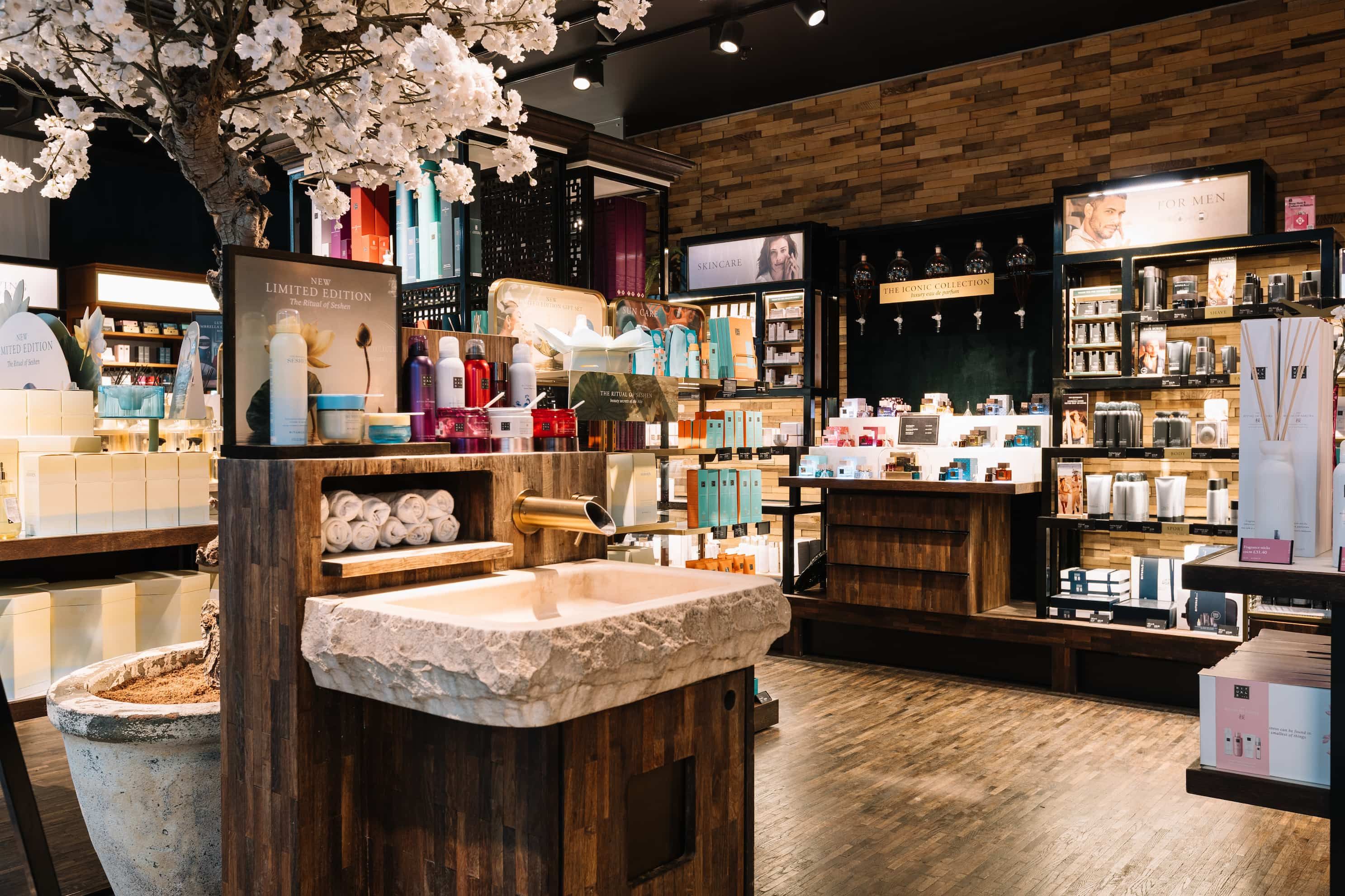 Luxurious skincare store with wooden décor, a stone sink, flower arrangements, and well-organised shelves of products and towels.