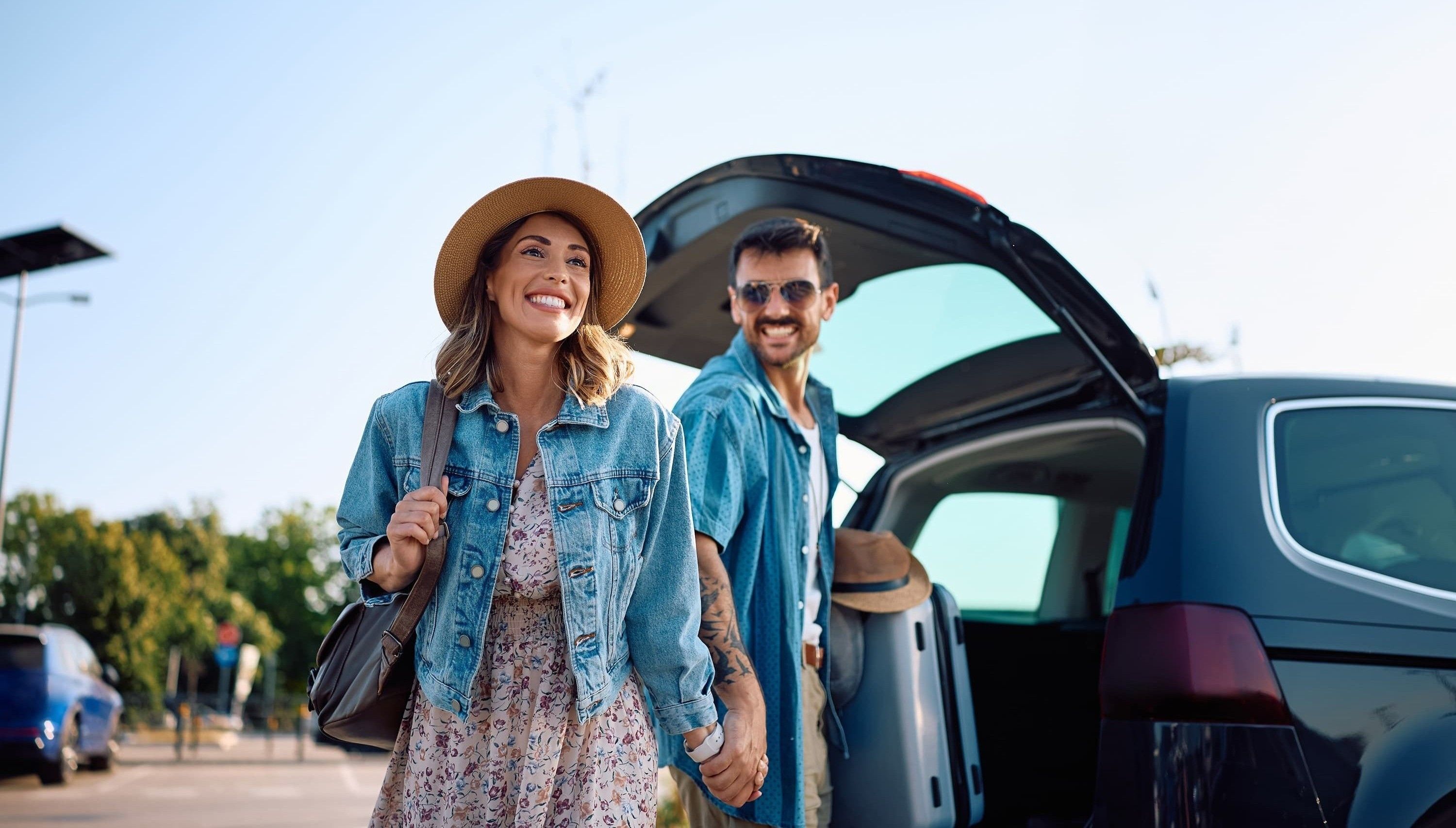 A smiling couple in casual clothes holds hands near an open car trunk on a sunny day, with trees and a clear sky in the background.