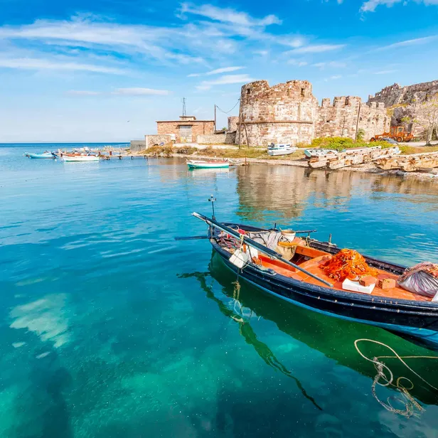 A small fishing boat floats in clear turquoise water near a rustic stone fortress on a bright day with blue skies.