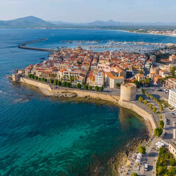 Aerial view of a coastal town with red-tiled roofs, a fortress, and a marina, surrounded by clear blue sea and distant hills.