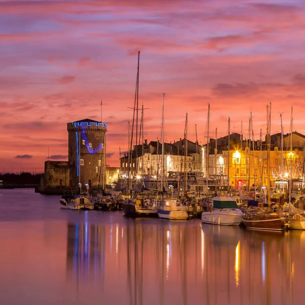 Harbor at sunset with two illuminated towers, docked sailboats, and a vibrant sky reflecting on the water, creating a serene ambiance.