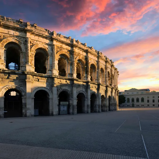 Ancient Roman amphitheater under a colorful sunset sky, with a spire and building in the background, set in an open plaza.
