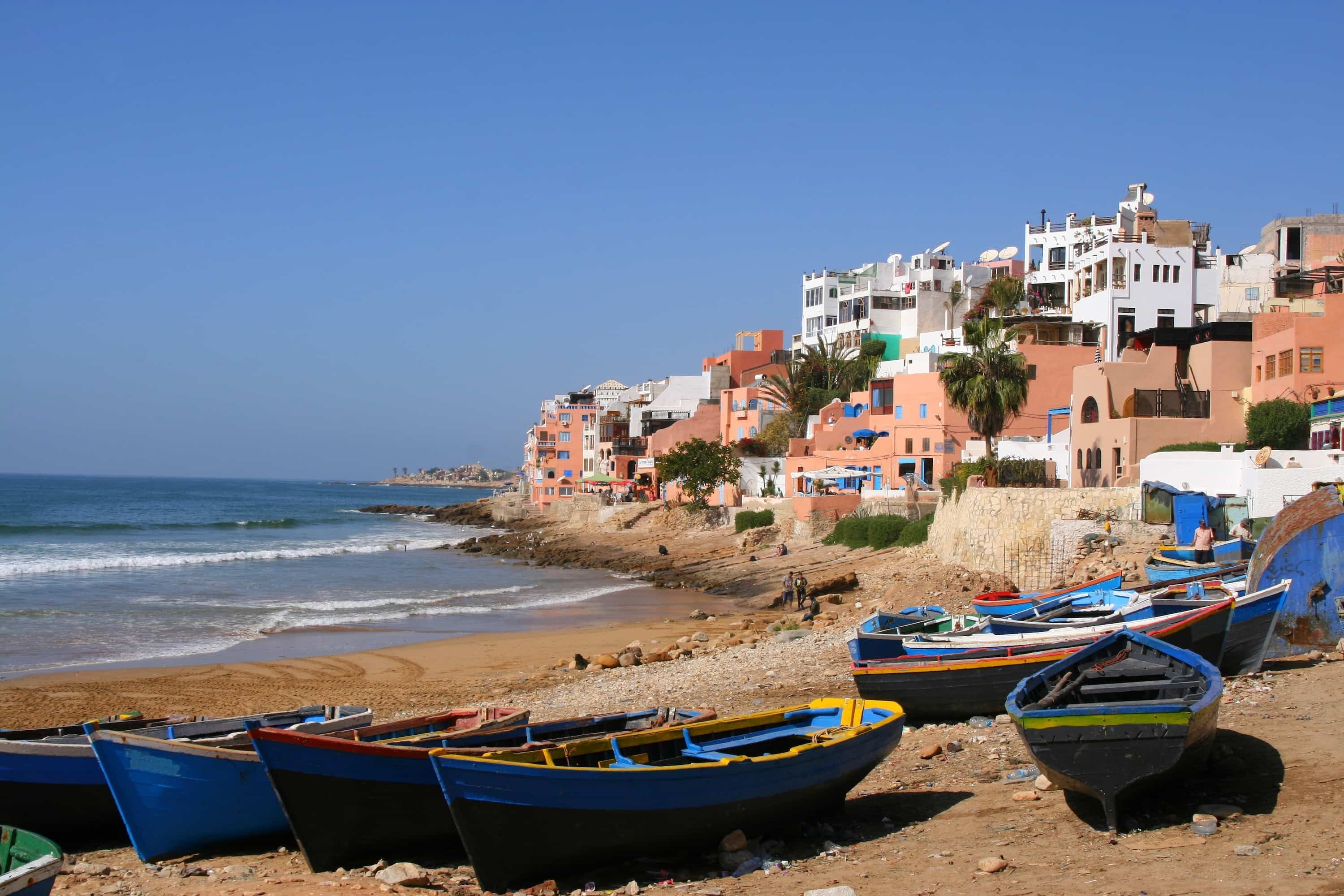 Colorful boats on a sandy beach with vibrant buildings on a hillside overlooking the ocean under a clear blue sky.