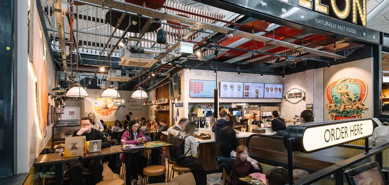 Interior of a bustling fast-food restaurant with passengers dining and ordering at the counter under a sign reading "LEON."