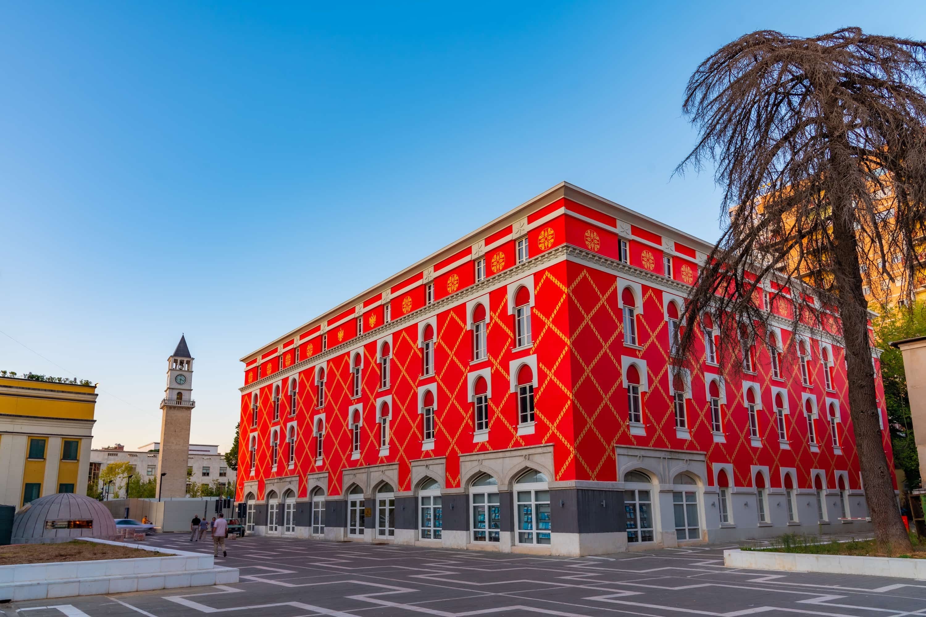 Vibrant red building with geometric patterns, arched windows, and a clock tower in the background under a clear blue sky.