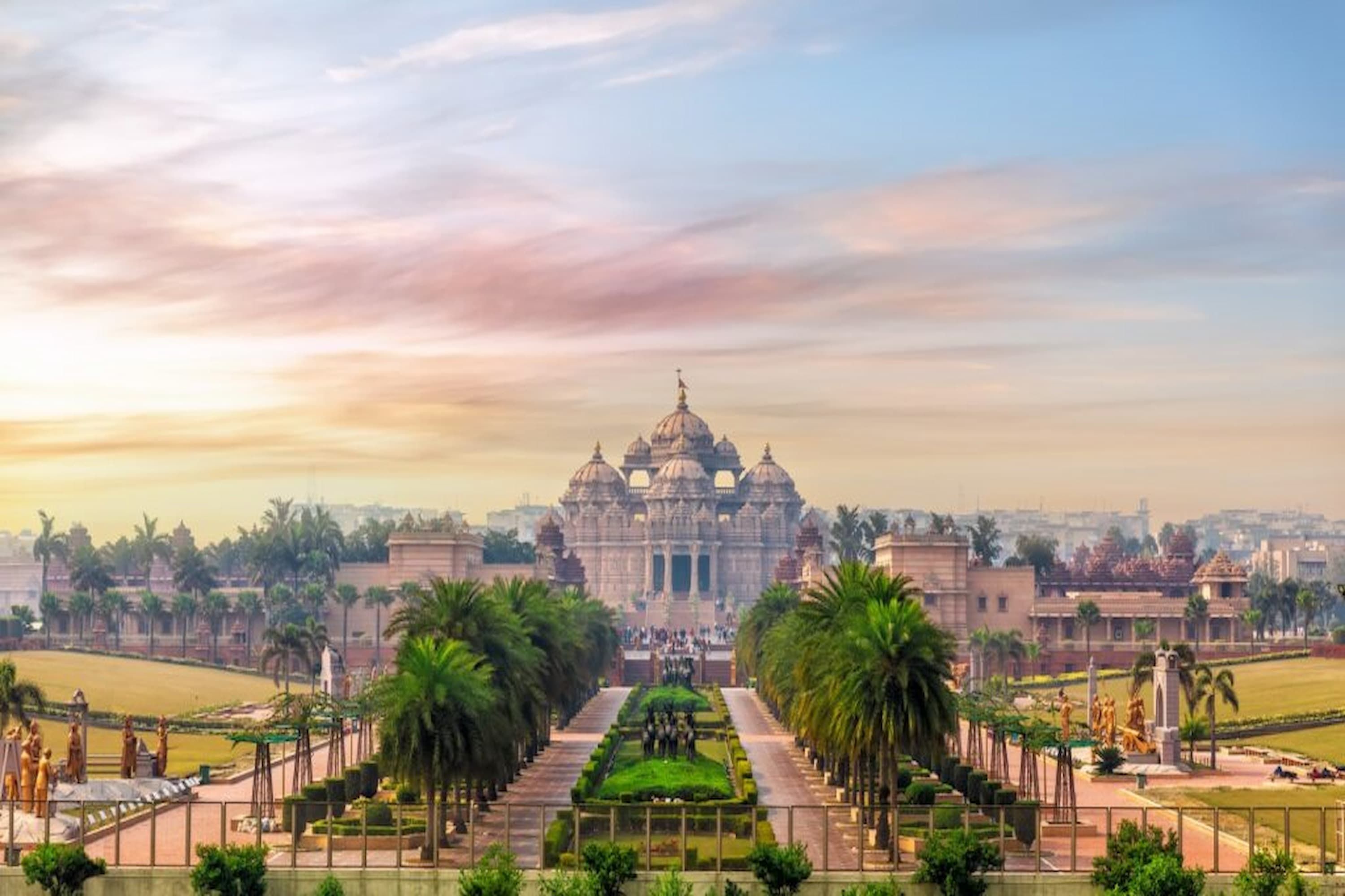 A grand temple complex with intricate architecture, surrounded by lush gardens and palm trees, under a colorful sky at sunset.