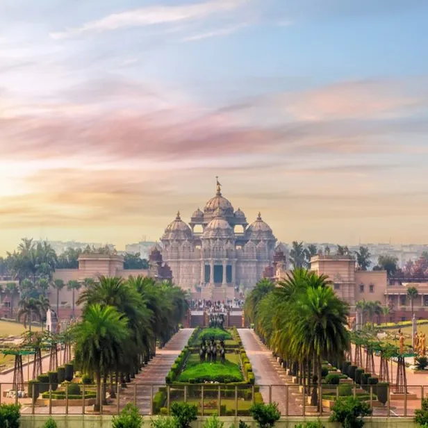 A grand temple complex with intricate architecture, surrounded by lush gardens and palm trees, under a colorful sky at sunset.