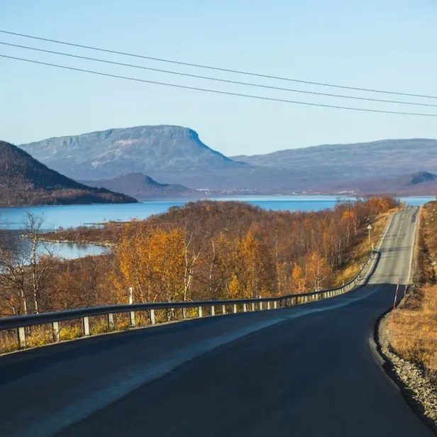 Scenic road with guardrails, flanked by autumn trees and hills, leads towards a tranquil lake and distant mountains under a clear blue sky.