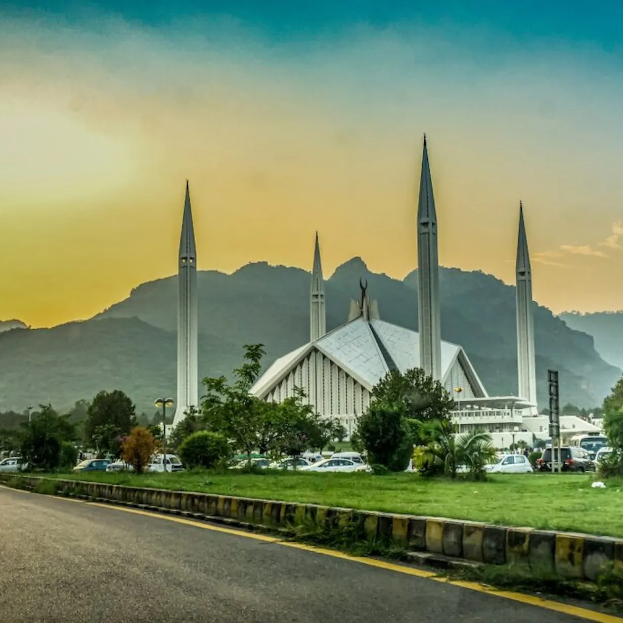 A mosque with tall minarets against a mountain backdrop at sunset, surrounded by greenery and parked cars.
