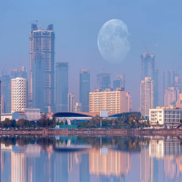 City skyline with modern skyscrapers at dawn, a large moon visible in the sky, and serene water reflections in the foreground.