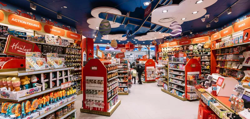 Colourful toy store interior with aisles of action figures, vehicles, and soft toys under a decorative ceiling featuring clouds and a plane.