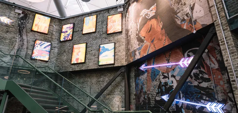 Modern interior with a brick wall featuring graffiti art, neon arrows, and vibrant framed posters. A glass staircase leads upwards.