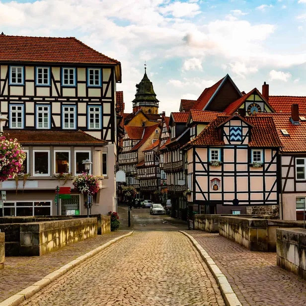 Cobblestone street leading to traditional half-timbered houses with red roofs, adorned with hanging flowers and a tower in the background.