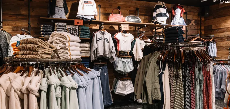 Clothing store interior with hoodies, sweaters, shorts, and jackets displayed on racks against a wooden wall, under bright lighting.