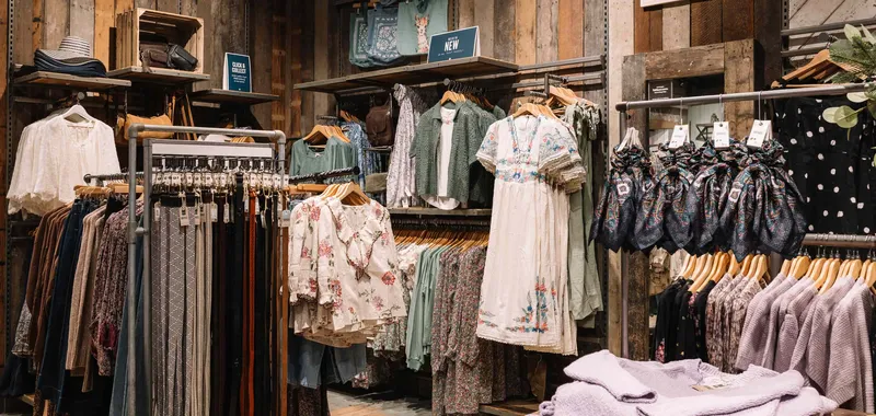 Clothing store interior with floral and patterned dresses, scarves, and display of pastel sweaters on rustic wooden shelves.
