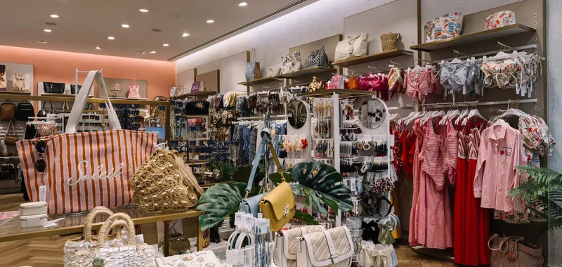 Clothing store interior featuring racks of colourful bags, dresses, jewellery, and accessories on display, with bright lighting and wooden flooring.