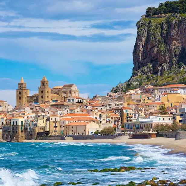 A picturesque coastal town with historic buildings and a cathedral, set against a rocky cliff and a clear blue sky, with waves crashing on the shore.