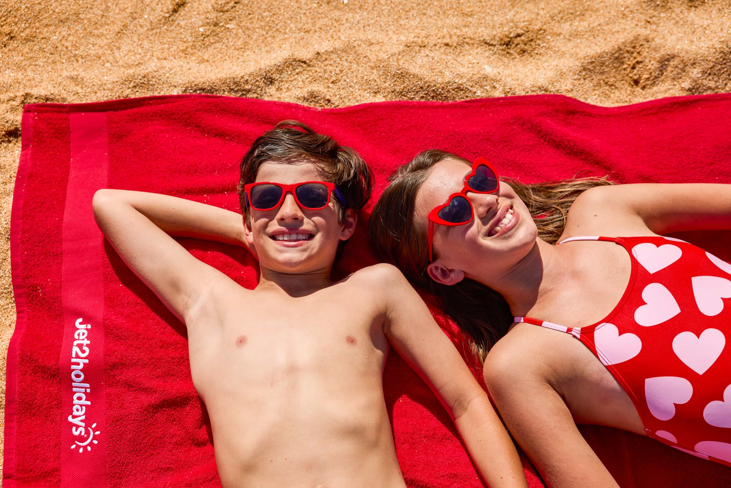 Two kids wearing red sunglasses sunbathe on a red beach towel with heart designs, smiling and enjoying the sunny day.