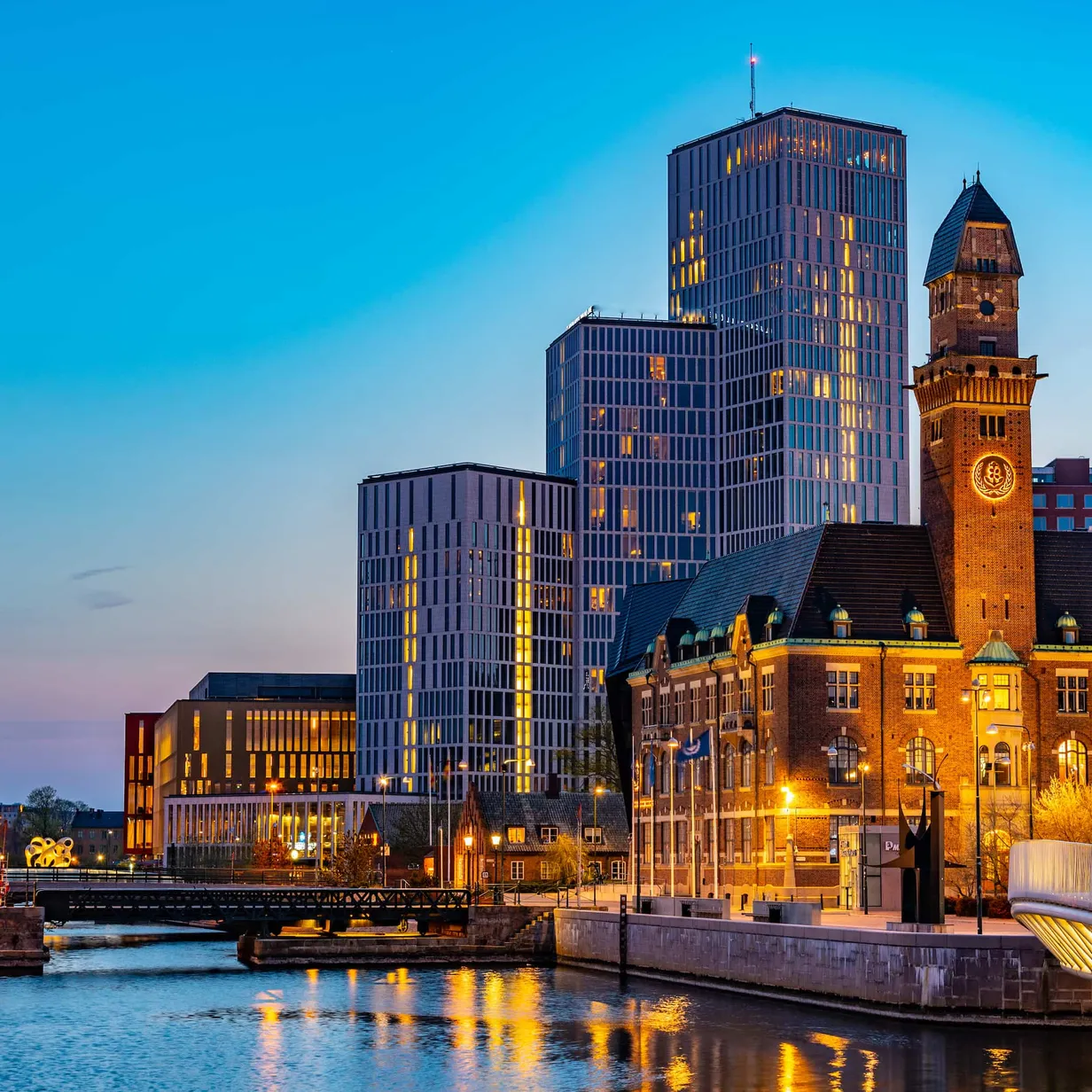 Cityscape at dusk with illuminated modern skyscrapers and historic buildings reflected in a canal.