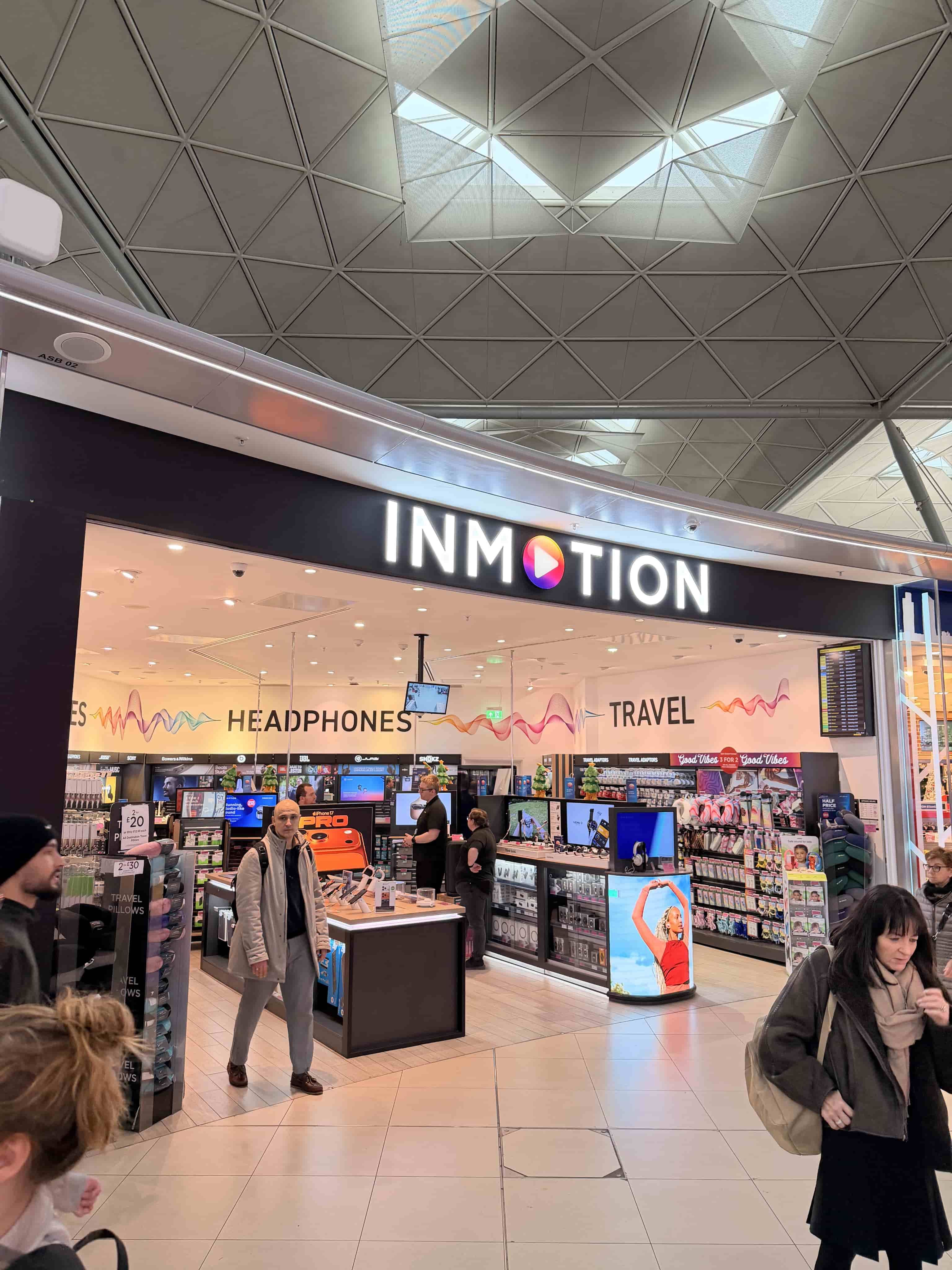 Airport electronics store INMOTION with illuminated sign, headphones and travel gear displays, shoppers walking by.