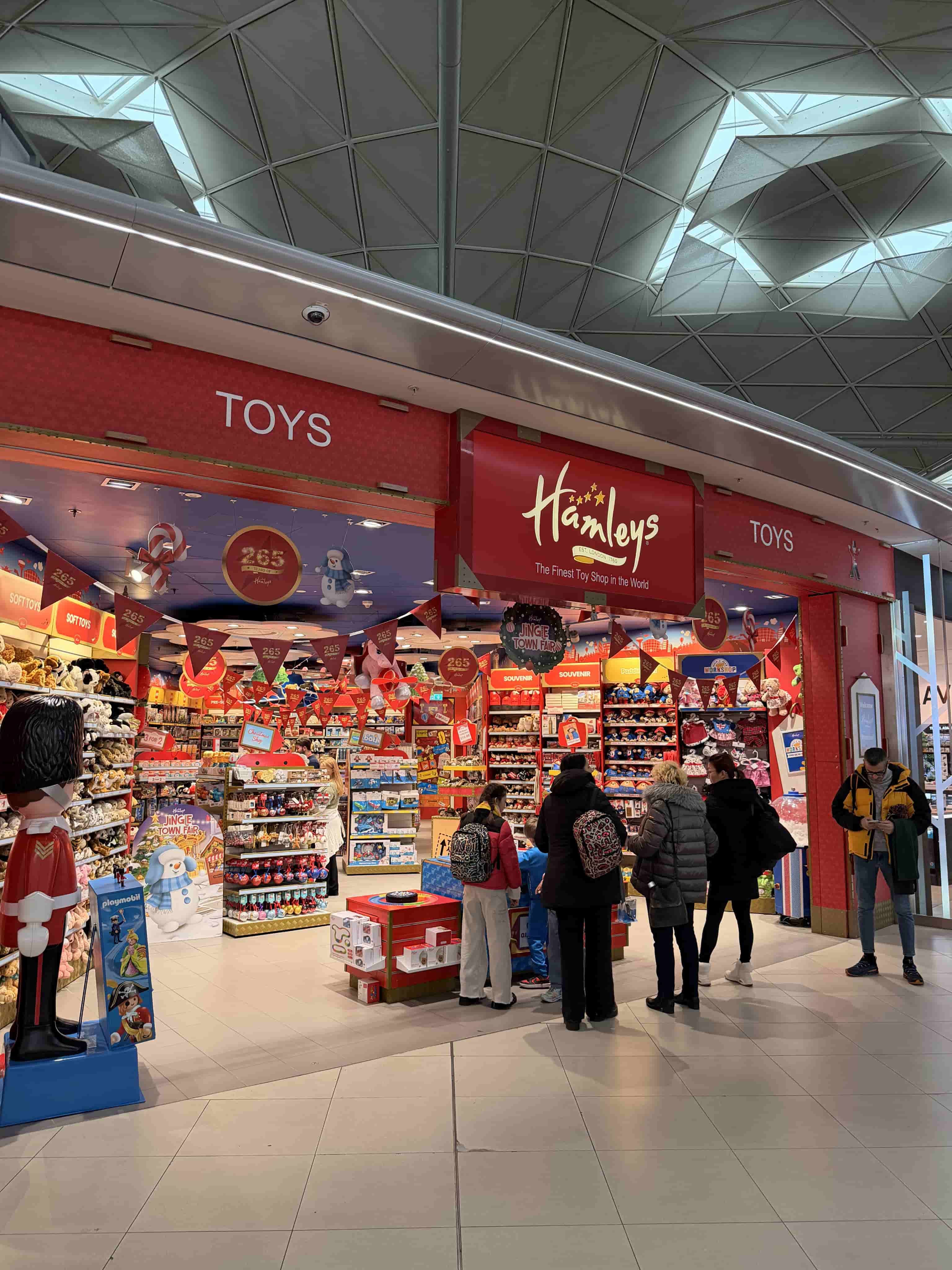 Hamleys toy store entrance in a mall, red signage, crowded shelves and shoppers browsing toys.