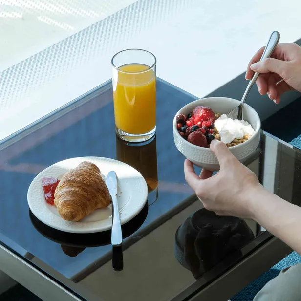 A person holds a bowl of granola with yogurt and berries beside a glass of orange juice and a plate with a croissant and jam on a glass table.