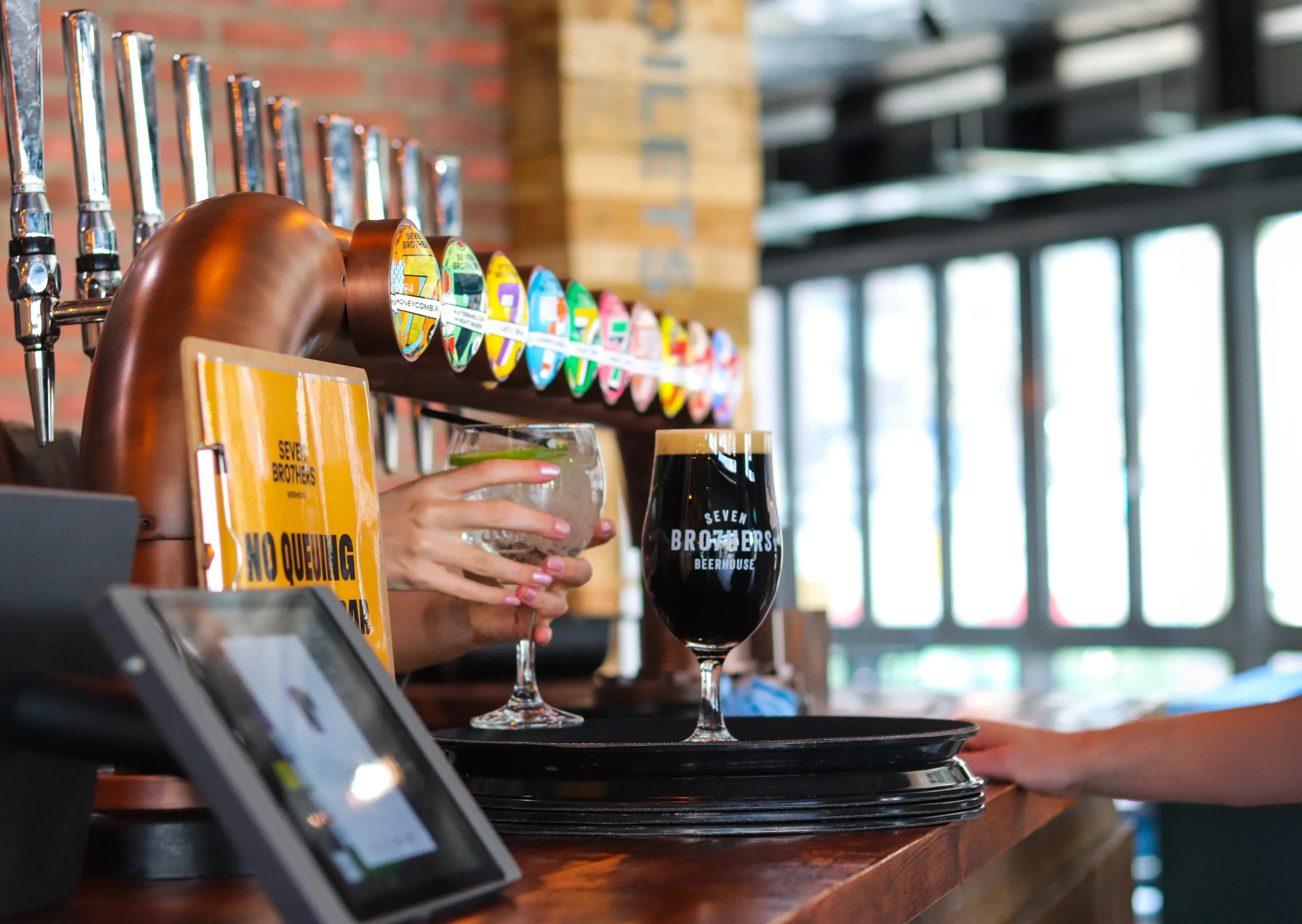 A person serves a cocktail and a dark beer on a tray at a bar, with colorful beer taps and a "No Queuing" sign in the background.