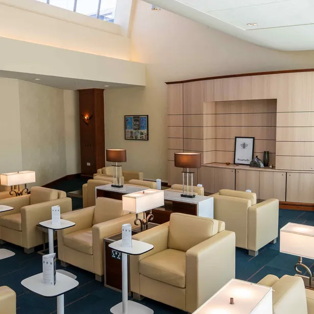 A modern lounge with beige armchairs, white side tables, and lamps. Wooden shelves and wall art decorate the room, creating a cozy ambiance.