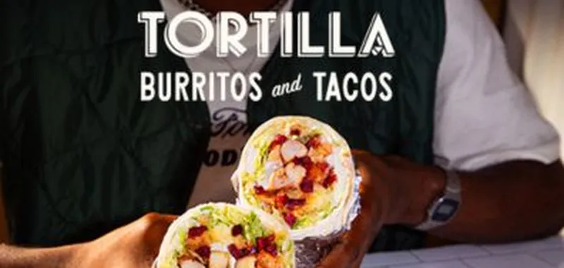 Person holding a halved burrito filled with ingredients, under a sign reading "Tortilla Burritos and Tacos."