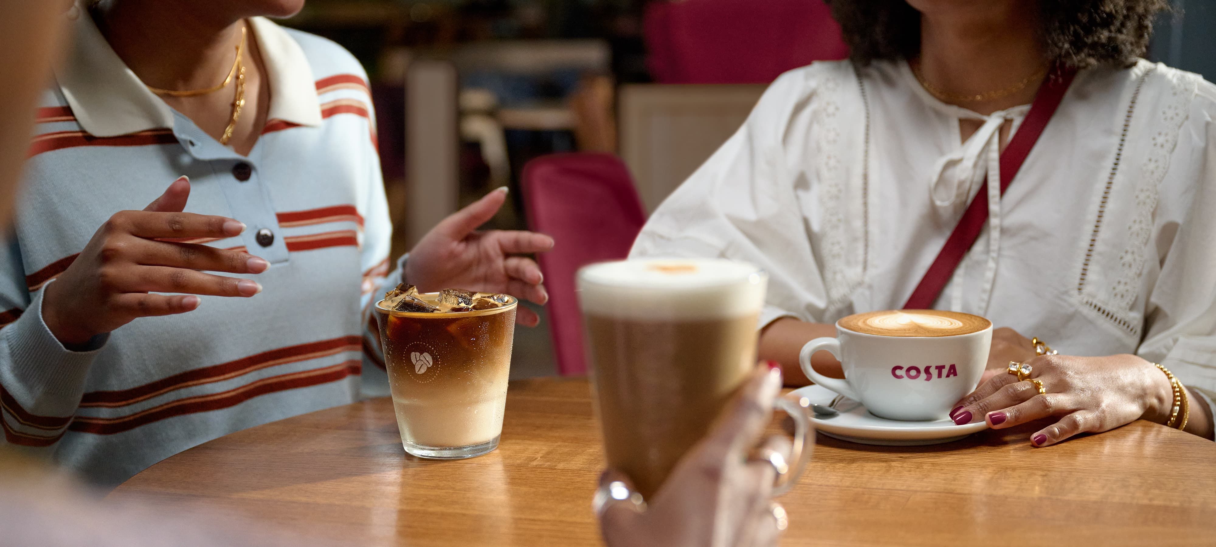 Three people at a café table, two with iced and hot coffee drinks, one with a latte in a Costa cup. They're engaged in conversation.
