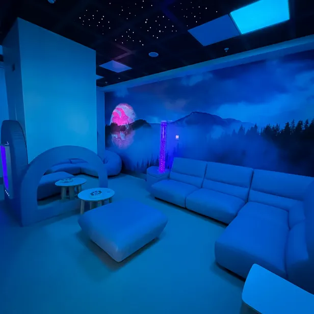 A blue-toned room with modern furniture, a starry ceiling, and a mural of mountains and a pink moon. Ambient lighting creates a calming atmosphere.