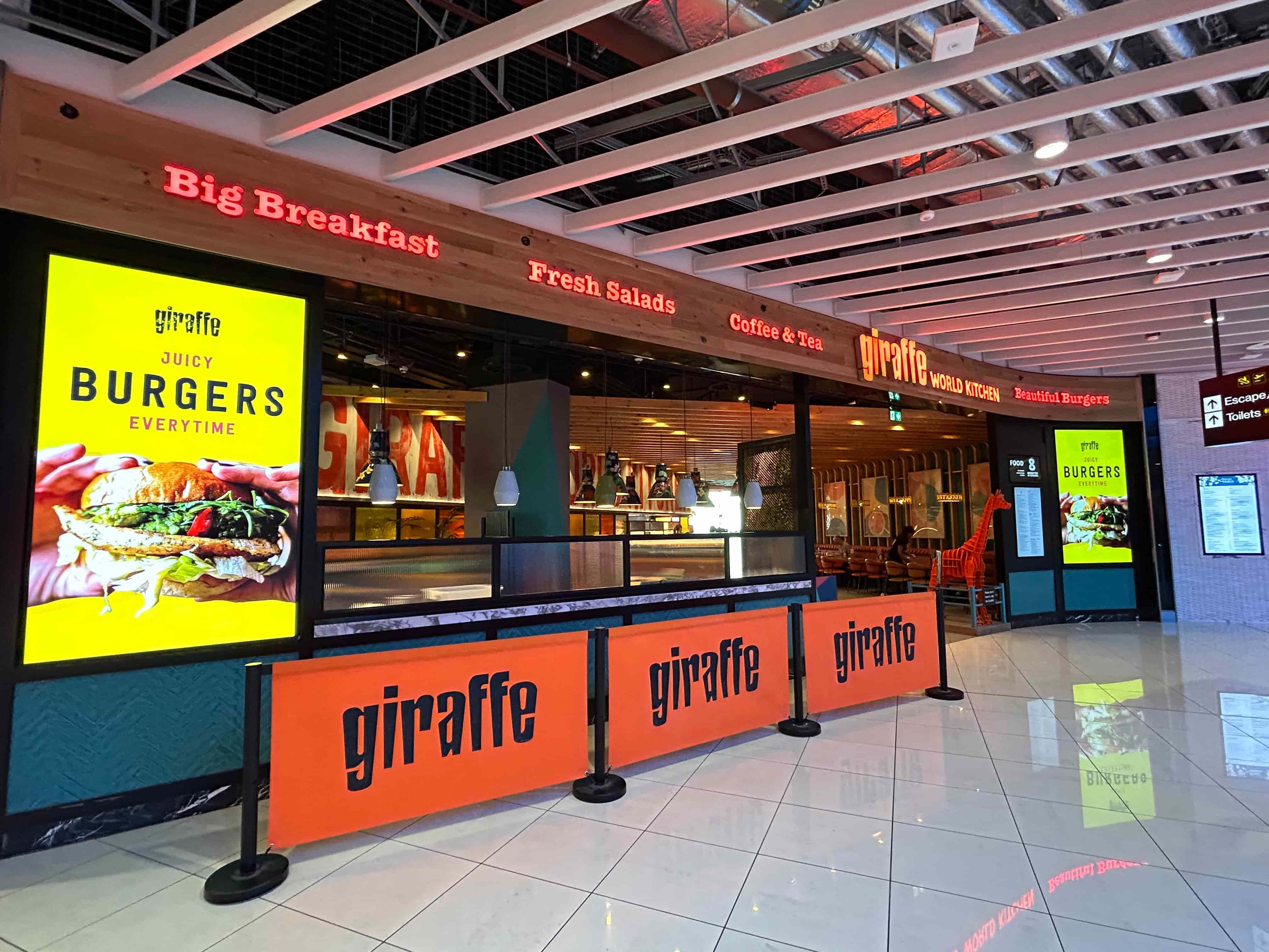 Front view of a Giraffe restaurant, featuring bright yellow burger advertisements, and a modern interior with a wooden ceiling.