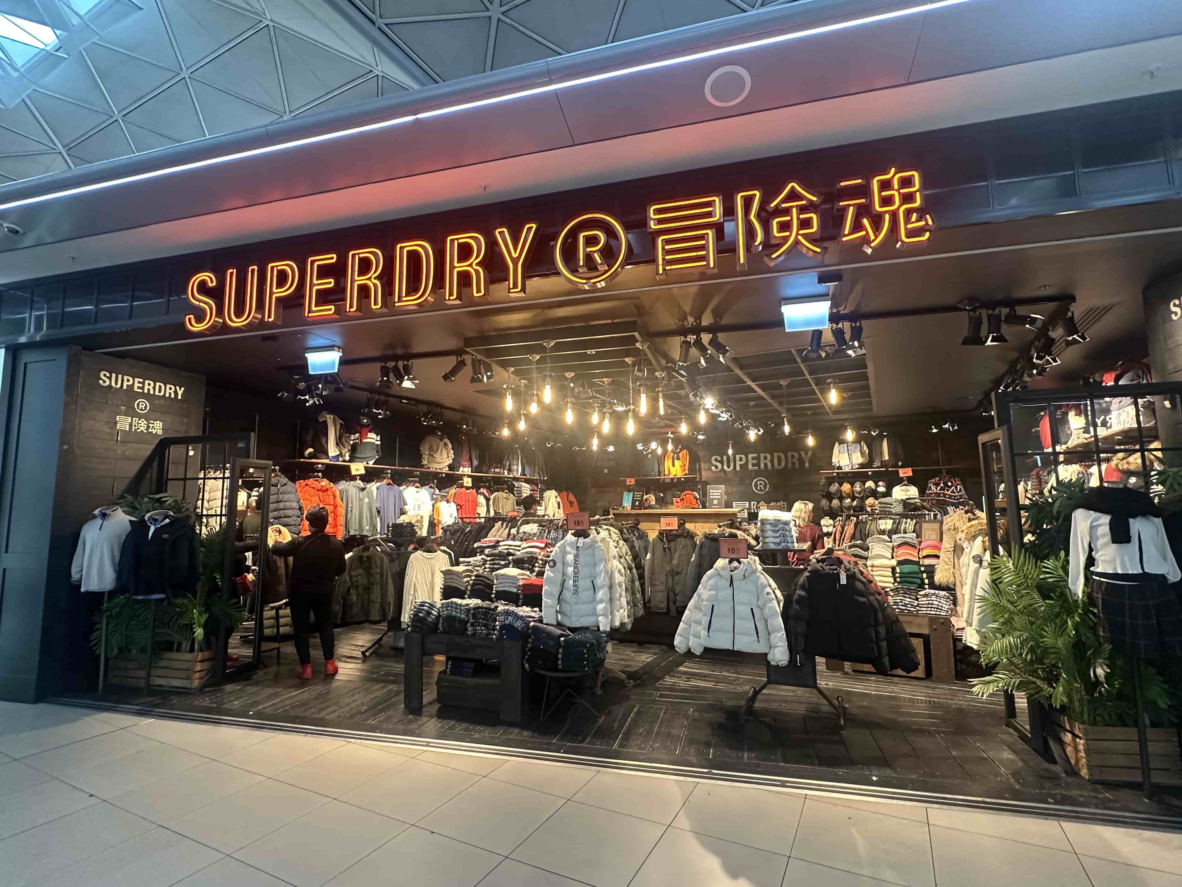 Superdry storefront with neon sign, lit interior displaying jackets, sweaters and racks, shopper browsing inside.