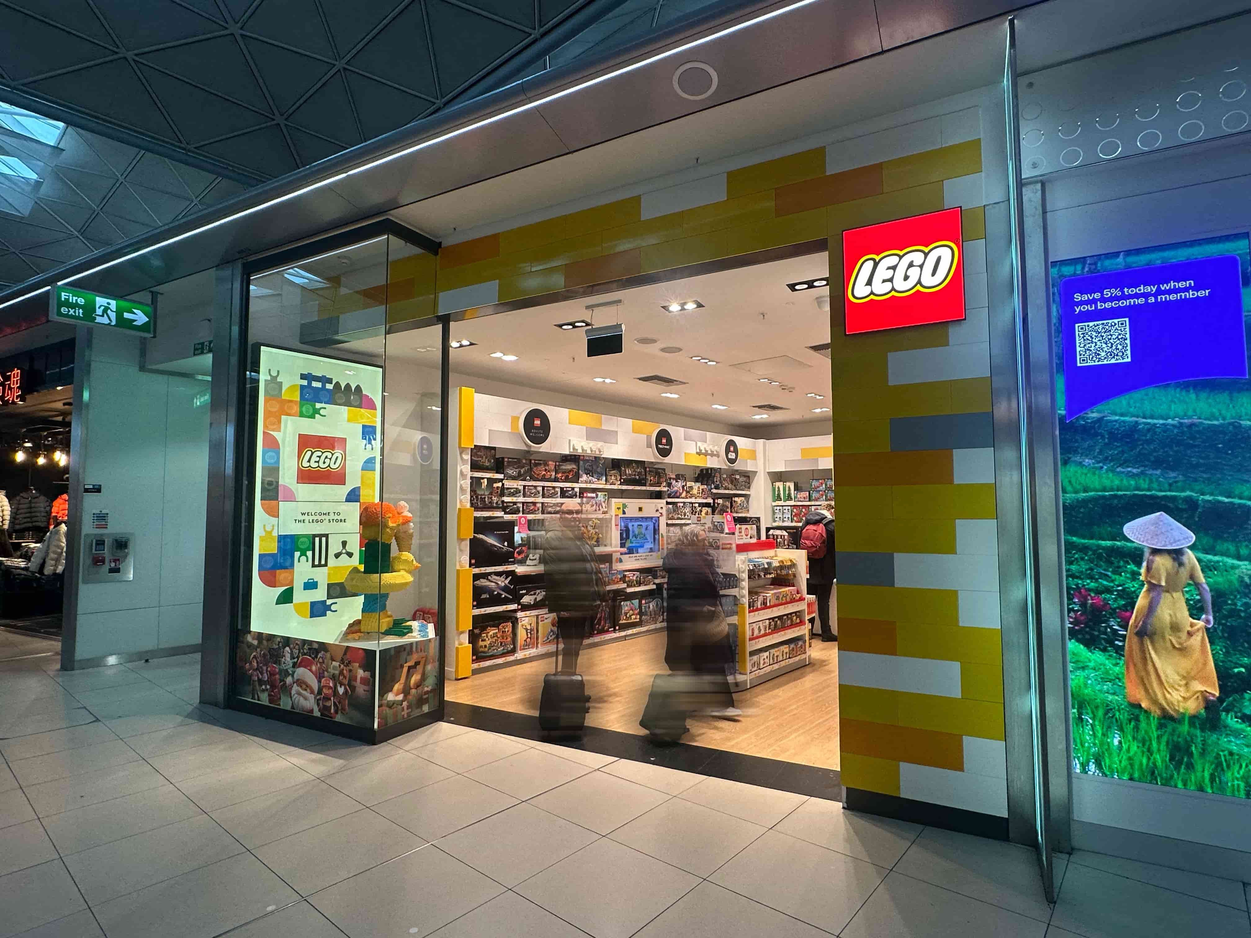 LEGO store storefront with illuminated red logo, yellow-and-white brick-pattern facade, window displays and blurred shoppers entering.