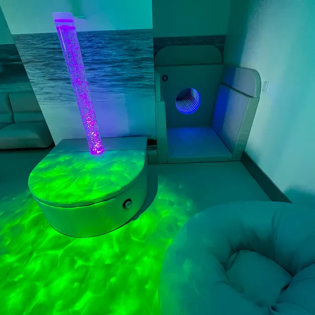 Room with a sensory bubble tube, green light reflections on the floor, cosy seating, and a small enclosed nook with blue lighting.