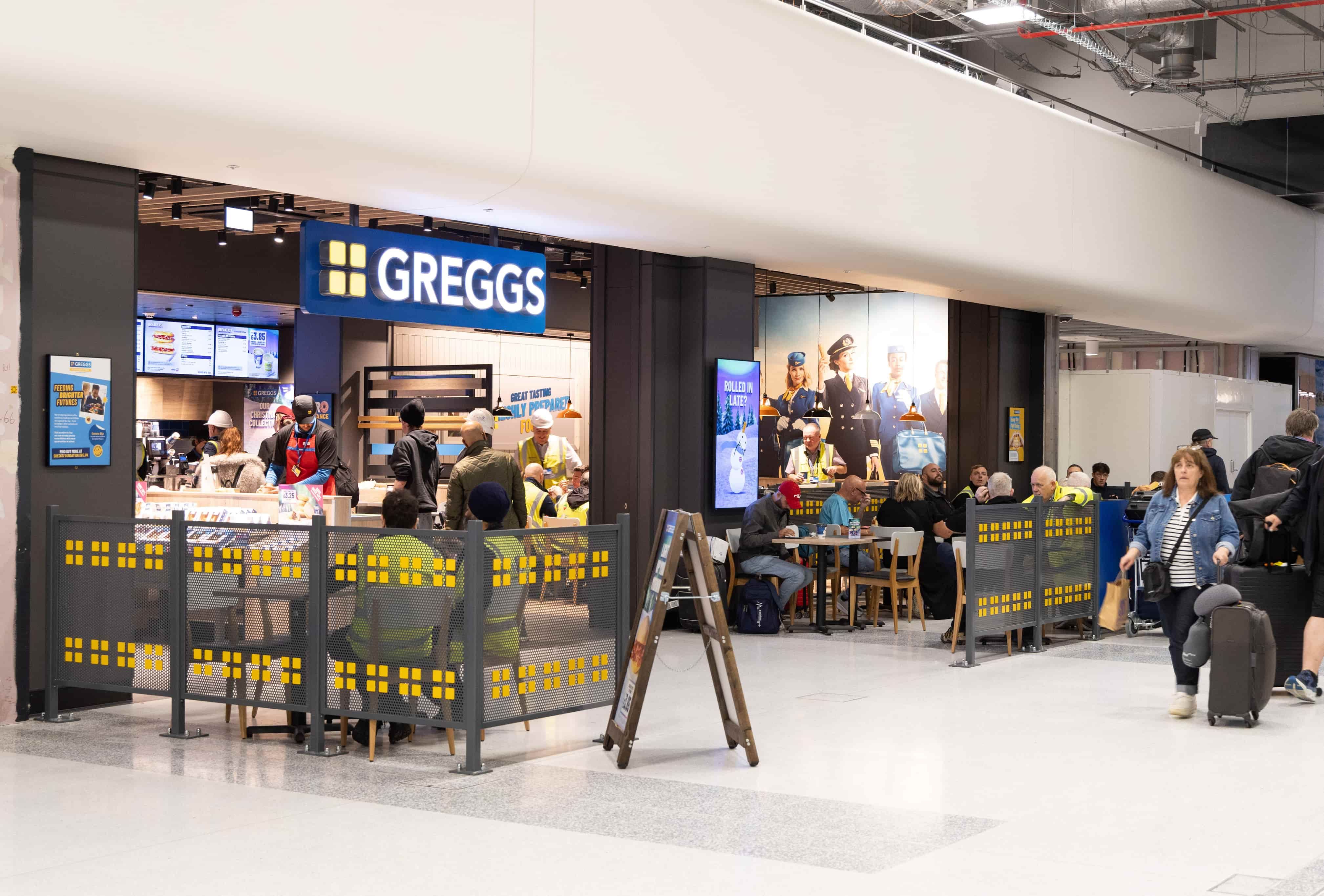 People seated and queuing at a Greggs outlet in an airport. Bright signage and surrounding seating area with travelers and luggage visible.