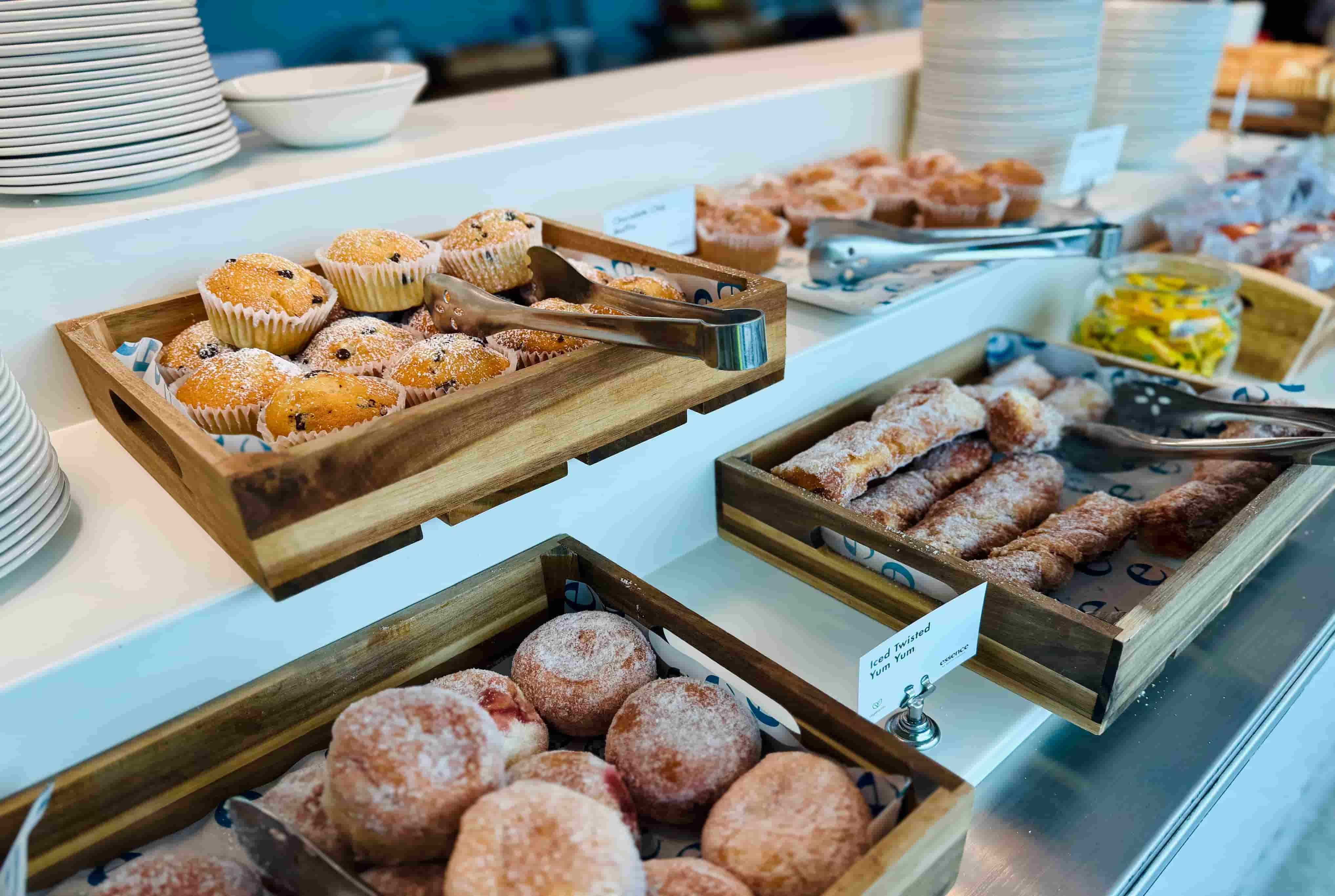 Assorted pastries, including muffins and donuts, displayed in wooden trays, with tongs and stacked plates in the background.