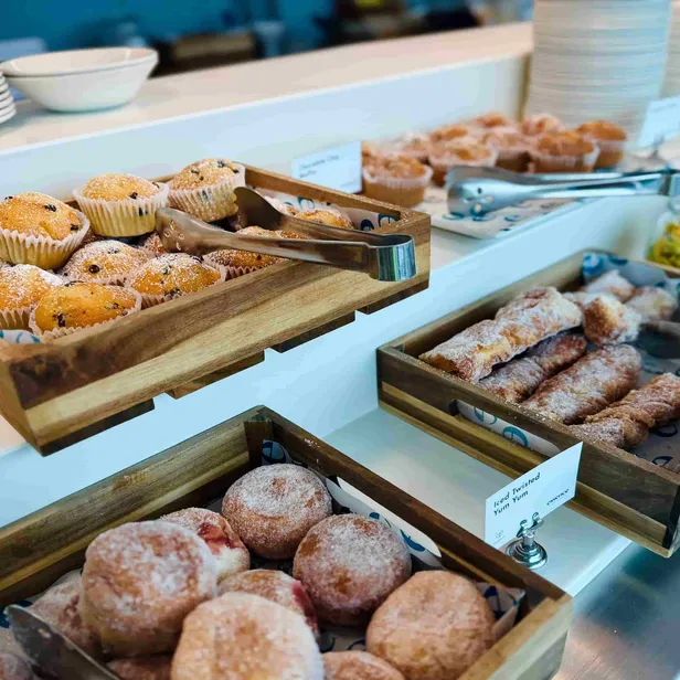 Assorted pastries, including muffins and donuts, displayed in wooden trays, with tongs and stacked plates in the background.