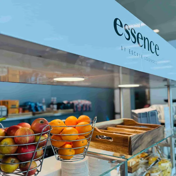 Fruits in wire baskets, cups, and cutlery on a buffet at a lounge labeled "essence by Escape Lounges."