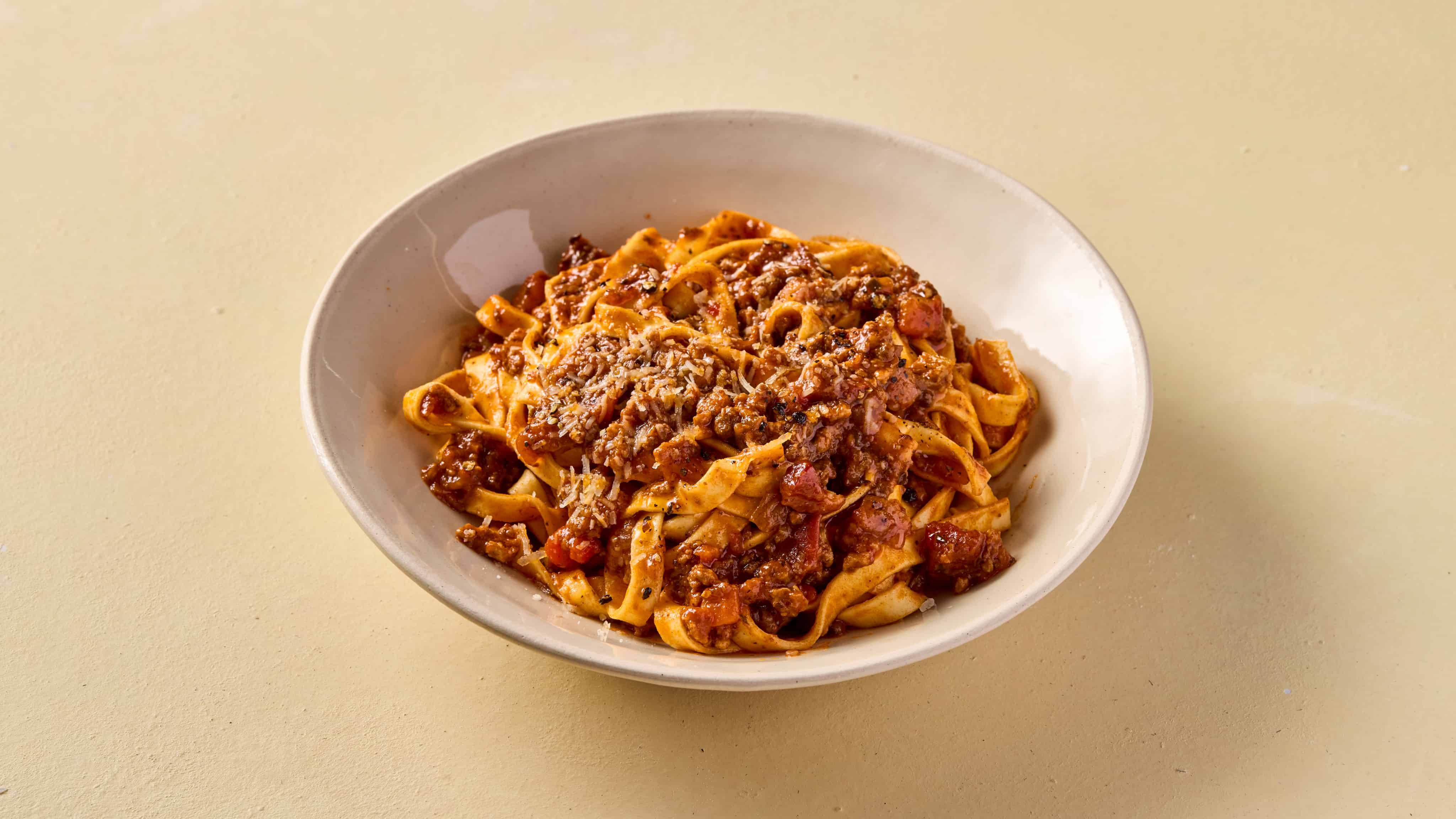 A bowl of tagliatelle pasta topped with a rich, meaty tomato sauce and sprinkled with grated cheese, set on a light-colored surface.