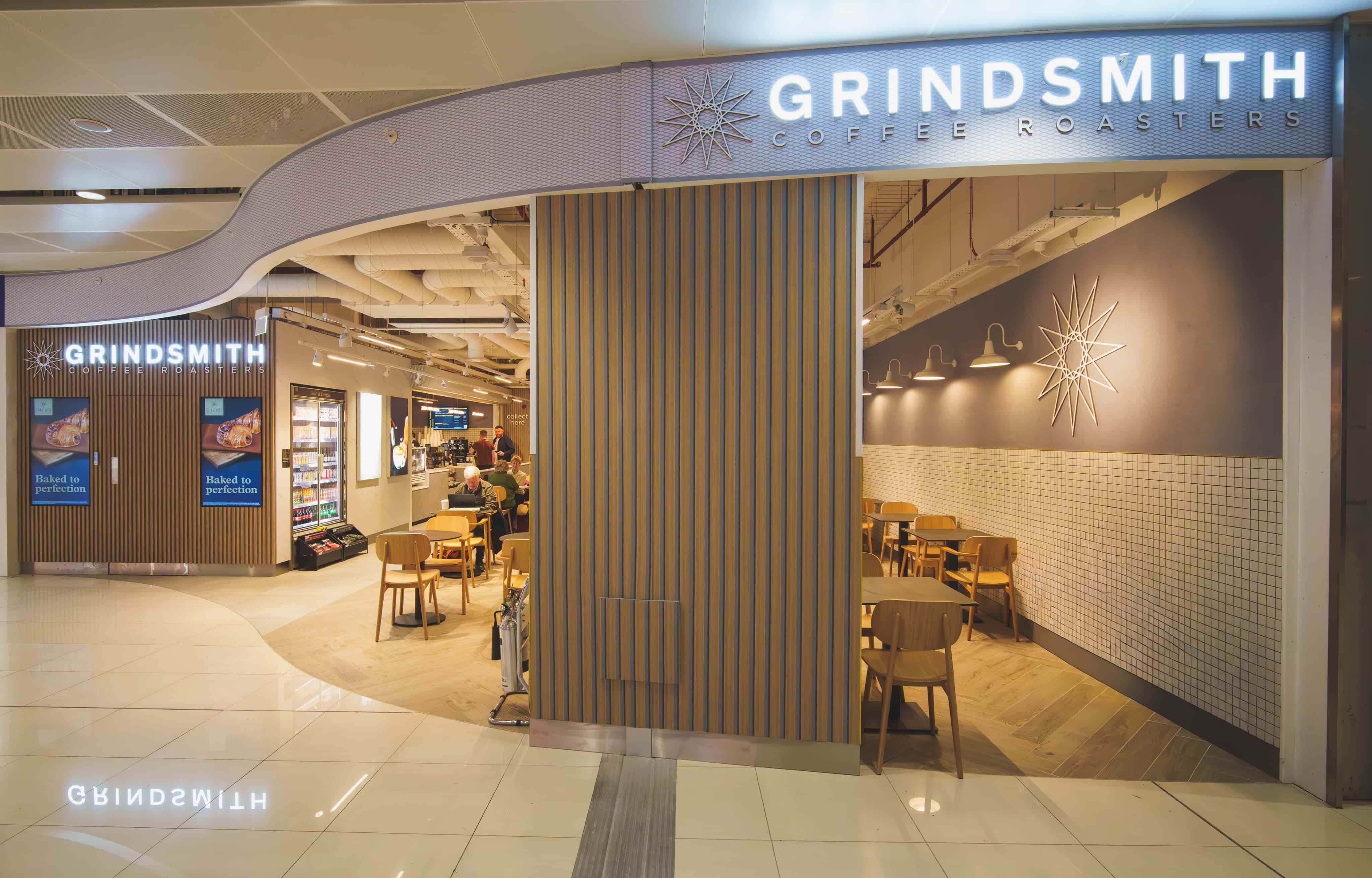 Cafe interior with a modern design, wooden tables, and chairs. "GRINDSMITH COFFEE ROASTERS" sign is visible above the entrance.