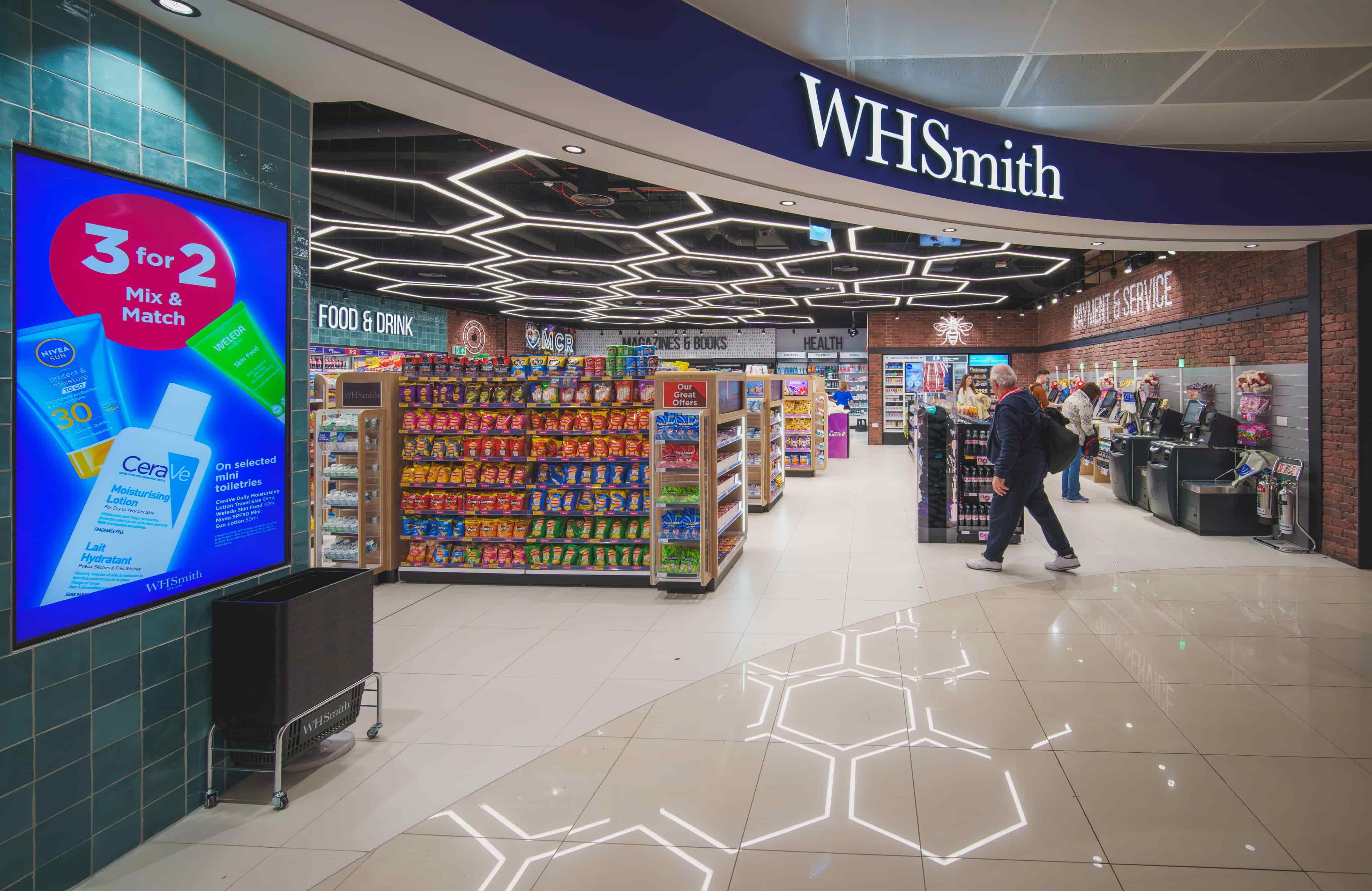 WHSmith store interior with snacks, books, and a blue "3 for 2" offer sign; geometric ceiling lights and customers shopping.