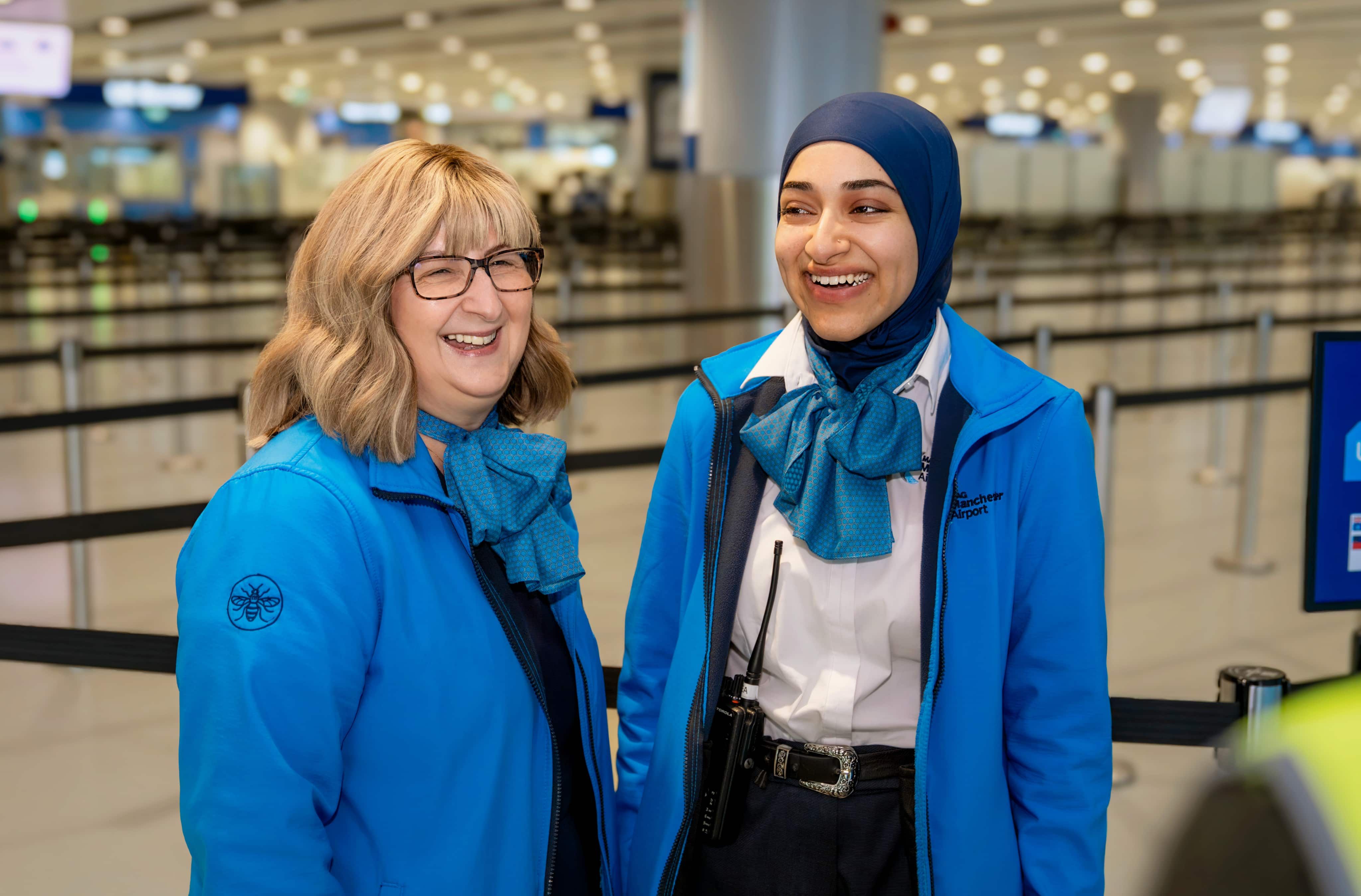 Two airport staff in blue uniforms, smiling and chatting at a terminal with blurred background.