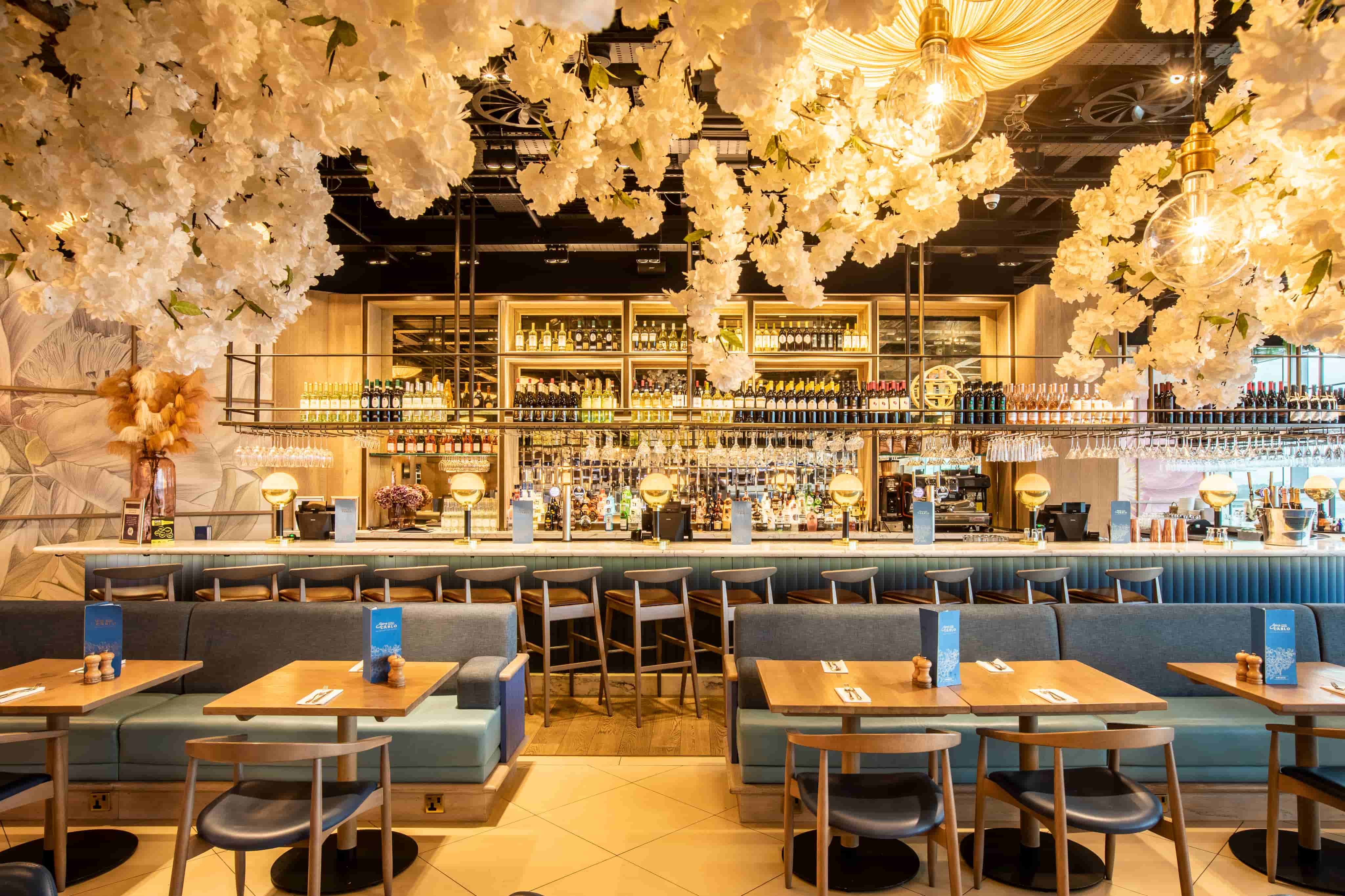 A stylish restaurant interior with floral ceiling décor, a fully stocked bar, blue cushioned seating, and wooden tables with menus.