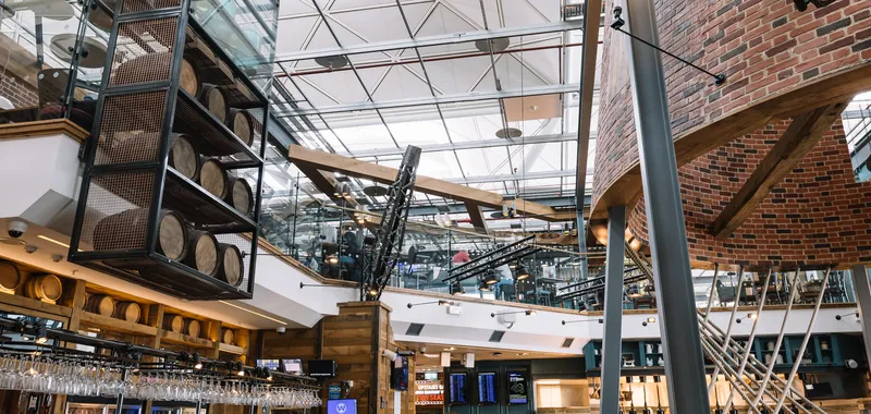 Modern, spacious bar interior with wooden barrels, glass panels, exposed beams, and a lively atmosphere. People are sitting and walking around.