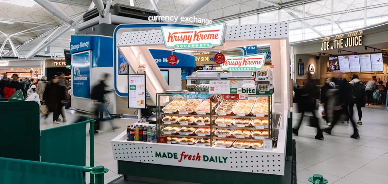 Krispy Kreme kiosk in a busy airport, displaying assorted doughnuts and drinks. Currency exchange and Joe & the Juice in the background.