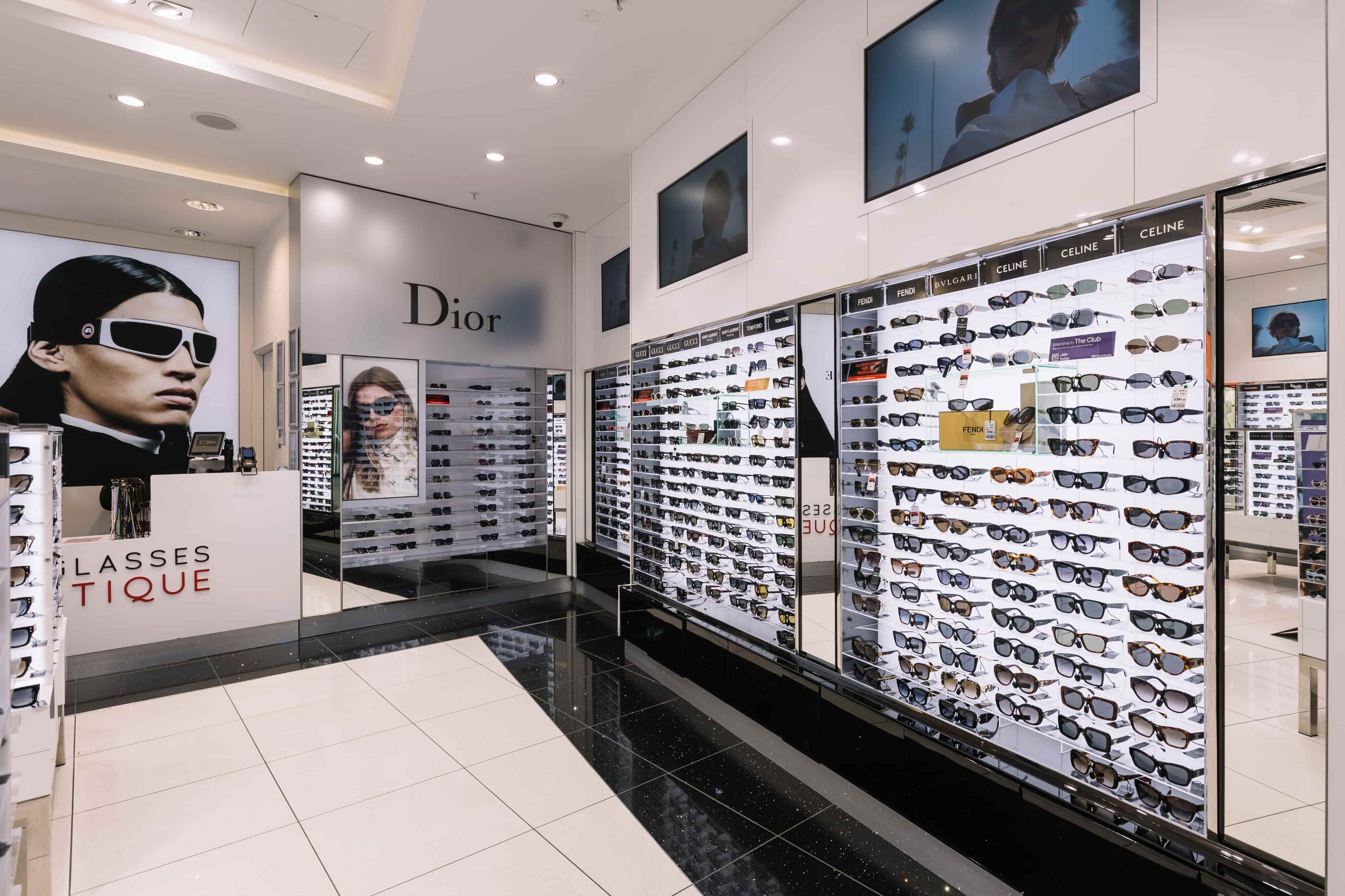 Sunglasses store interior with glass shelves displaying various designer brands like Dior, Gucci, and Celine, and large model posters.