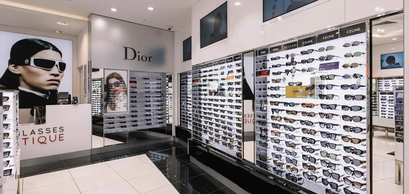 Sunglasses store interior with glass shelves displaying various designer brands like Dior, Gucci, and Celine, and large model posters.