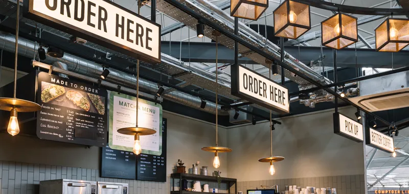 Cafe interior with signs reading "Order Here" and "Pick Up," hanging lights, menu boards, and a counter with kitchen equipment and cups.