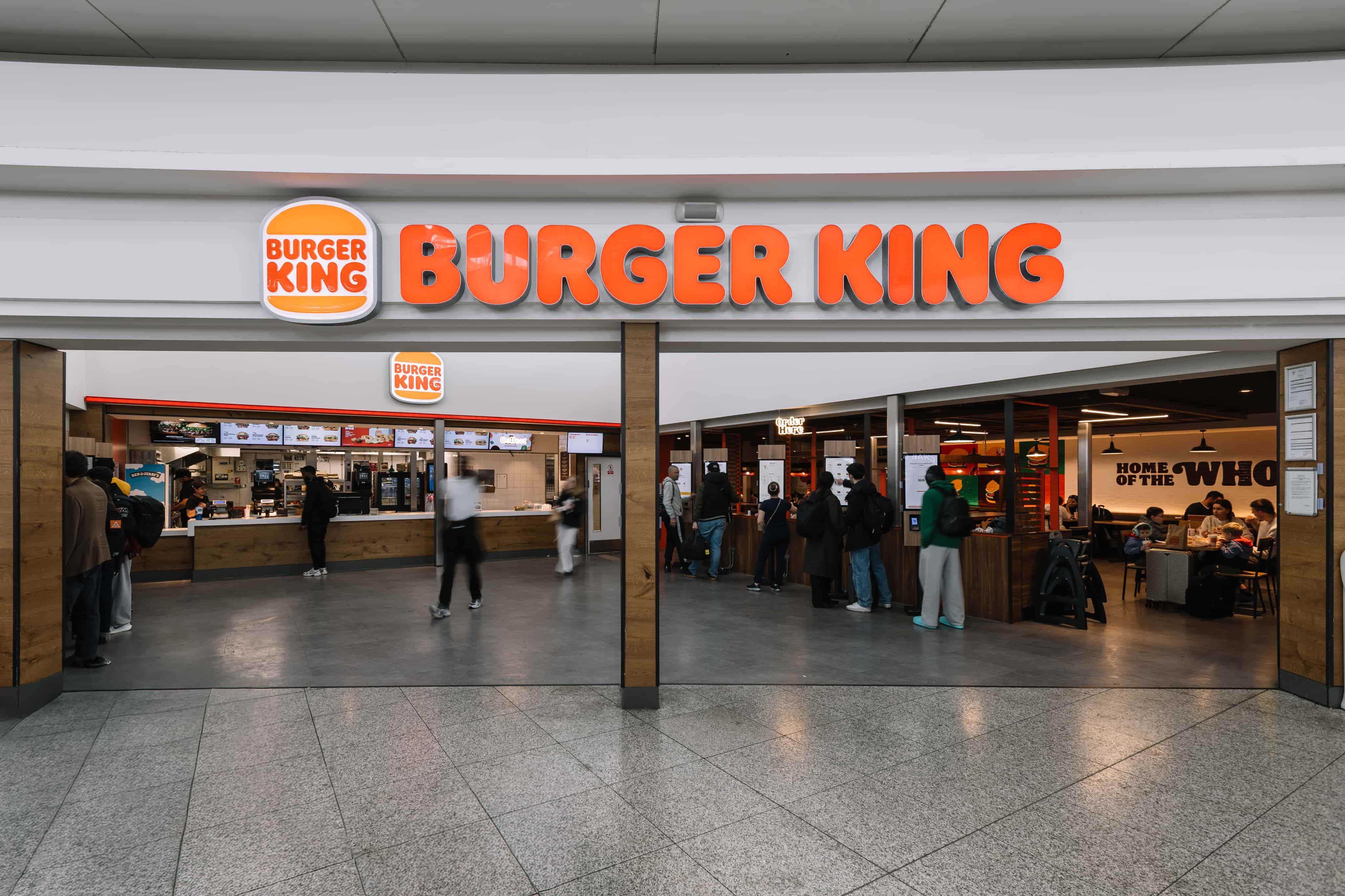 People standing in line at a Burger King restaurant inside a modern building; customers can be seen dining inside.